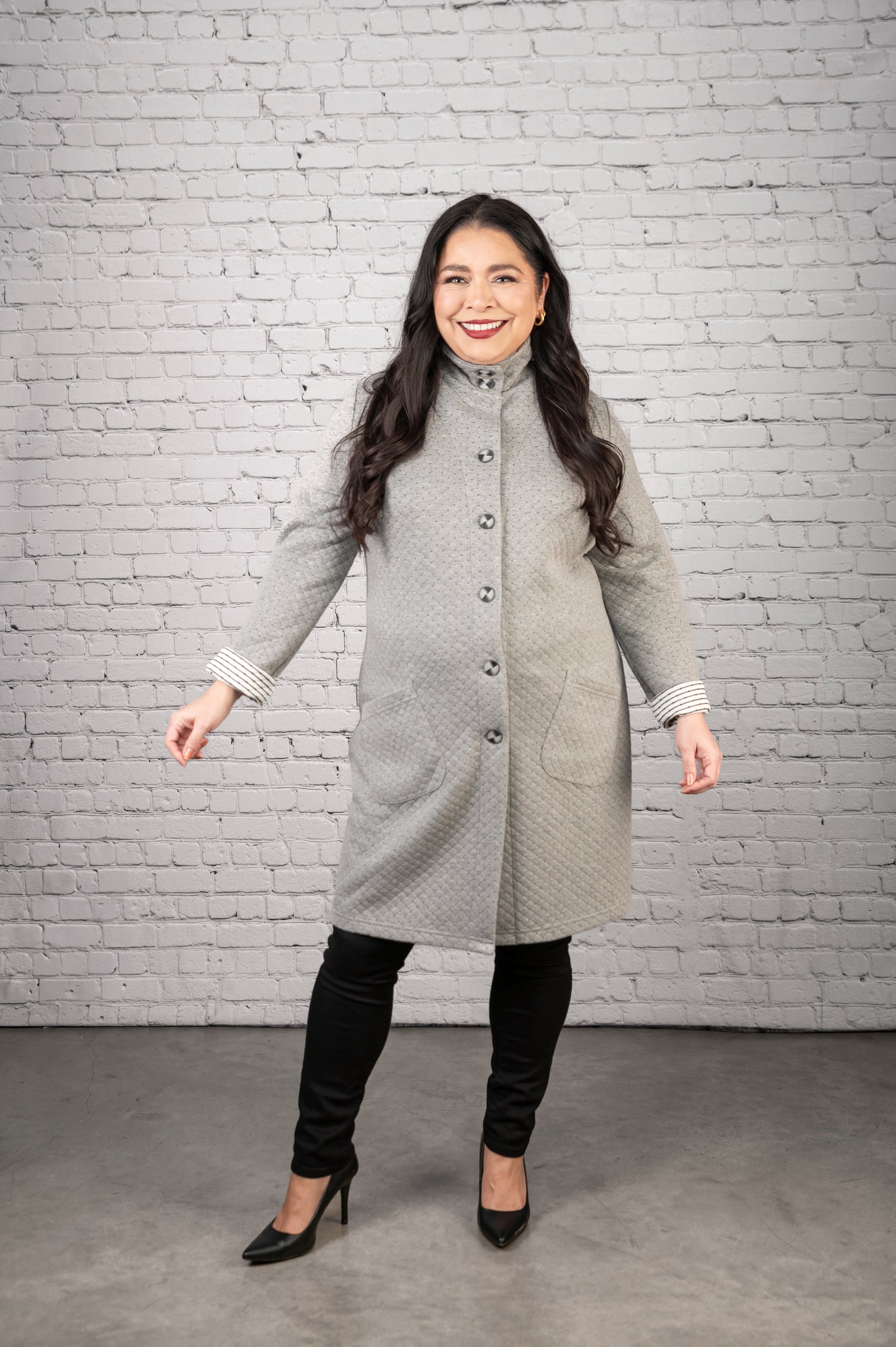 Woman wearing a gray coat standing against a white brick wall.