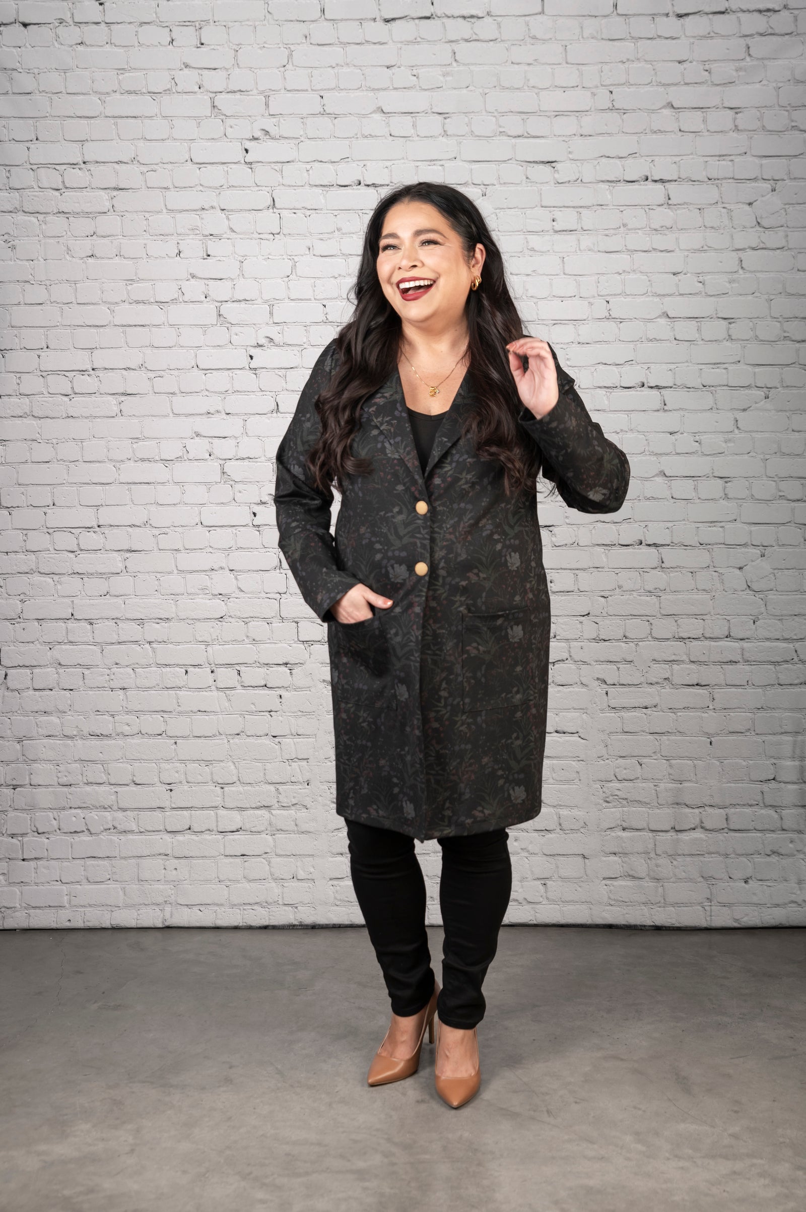 Woman wearing a dark gray coat standing against a white brick wall.