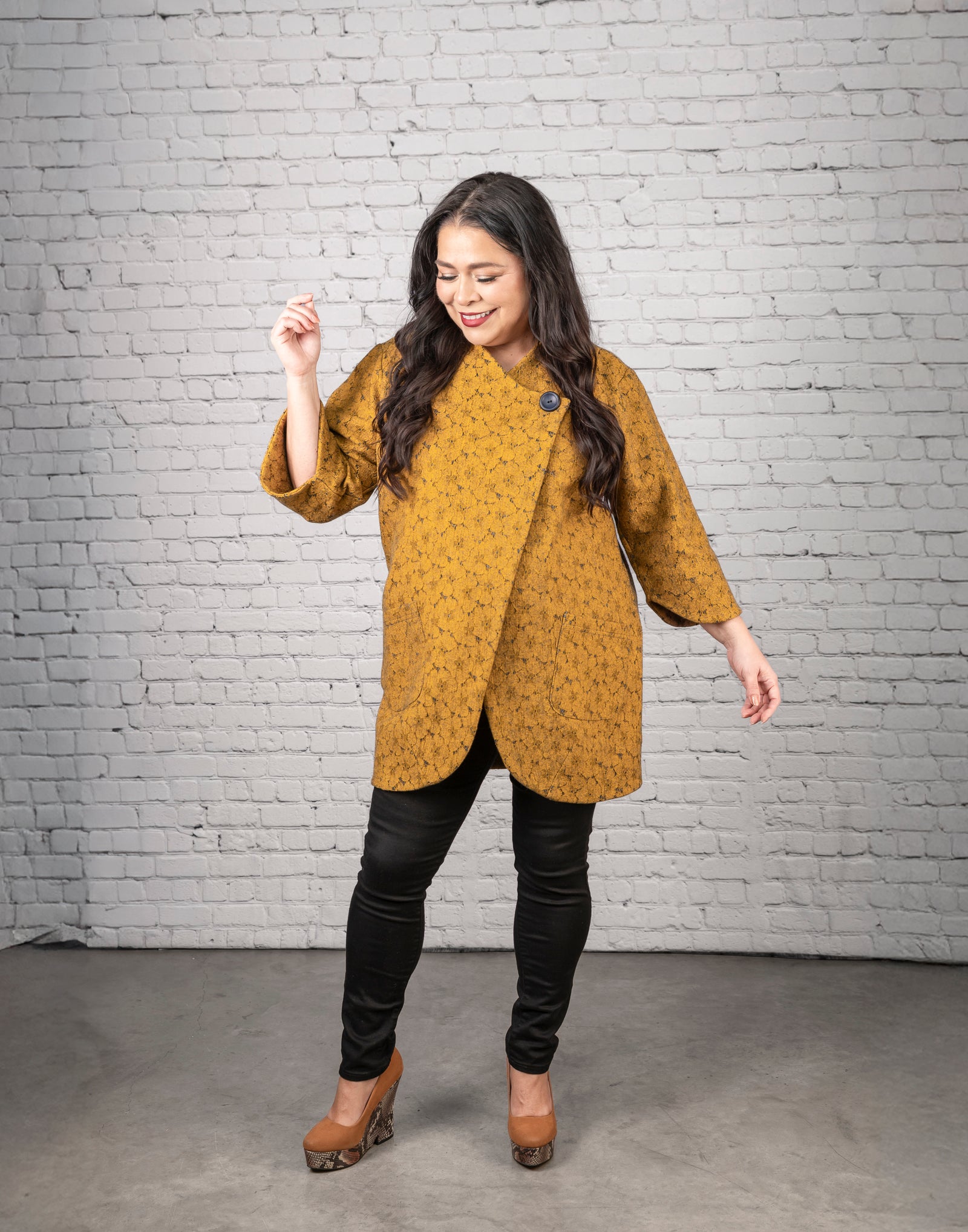 Woman wearing a mustard yellow top and black pants against a white brick wall.